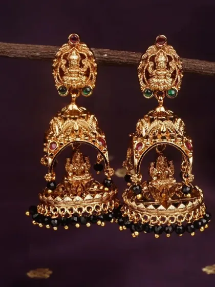 A close-up of the traditional temple jhumkis with a matte gold finish and black bead drops. The intricate detailing on the Lakshmi motif is clearly visible.