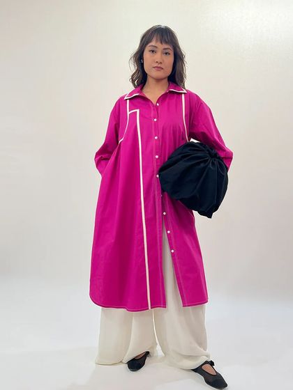 Draped in confidence, colored in power. A model wears a longline fuchsia shirt with white piping over wide-leg ivory trousers.