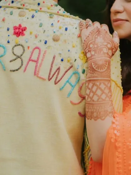 Forever and always. This intimate shot captures a beautiful, personalized detail on the groom's jacket, embroidered with a promise, as the bride rests her hand on his shoulder, showing off her intricate Mehendi design.