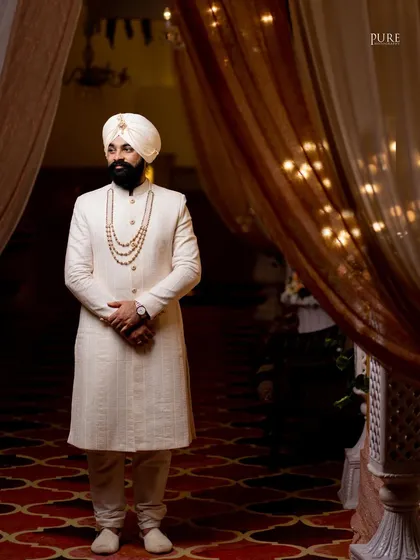 A striking portrait of the groom standing in a beautifully lit doorway. His elegant off-white sherwani and confident pose exude regal charm.