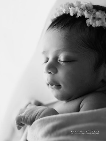 A black and white close-up of a newborn's face, with a delicate floral crown. The contrast highlights the softness of the baby and the texture of the flowers.