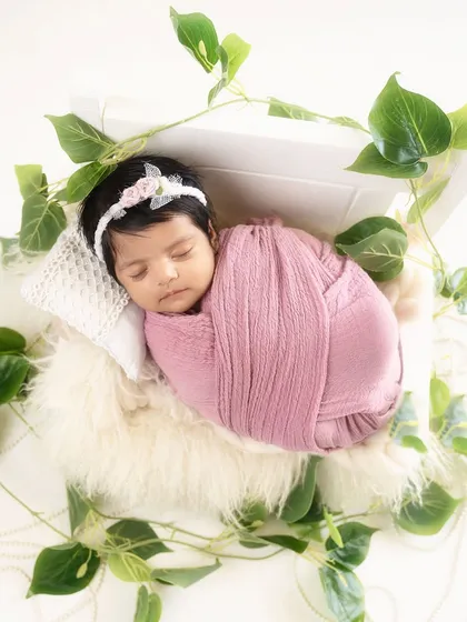 A dreamy portrait of a baby girl swaddled in pink, lying in a white box adorned with green vines. This setup is perfect for a magical, garden-themed newborn photo.