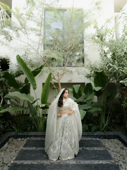 A full-length portrait of the bride in a stunning white lehenga, standing in a beautifully landscaped garden.