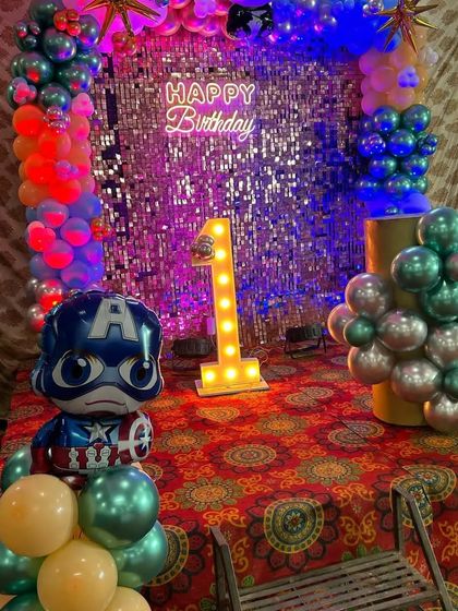 A superhero first birthday with a Captain America theme, a silver sequin wall, a colorful balloon arch, and a light-up number '1'.