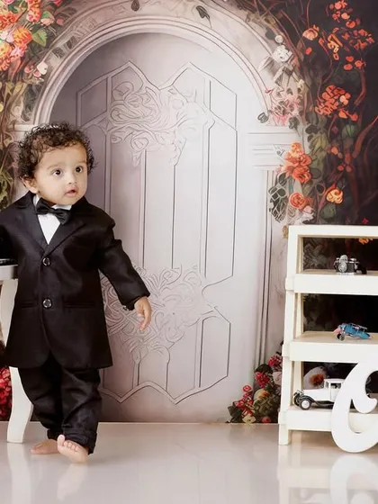 A dapper little gentleman in a tuxedo stands by a mini piano for his first birthday shoot, set against a floral backdrop.