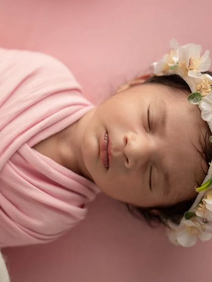 A delicate close-up shot of a newborn's face, wearing a floral crown and swaddled in soft pink fabric against a matching background.