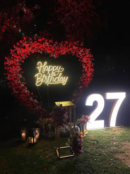 A romantic outdoor decoration featuring a large heart-shaped frame made of red flowers. This setup, shown here for a 27th birthday, is also perfect for a surprise proposal or anniversary celebration, complete with a custom neon sign and marquee numbers.