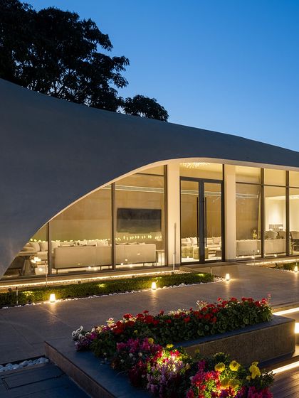 The full expanse of the glass facade of "The Rising Wave" glows warmly at night. The open-plan interior is visible from the garden, creating an inviting atmosphere for social gatherings.