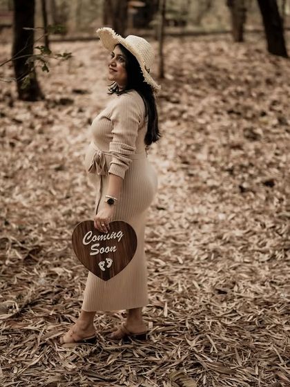 A lovely portrait of the mom-to-be in the woods, holding a "Coming Soon" sign and wearing a hat. The autumn leaves create a warm, rustic feel.