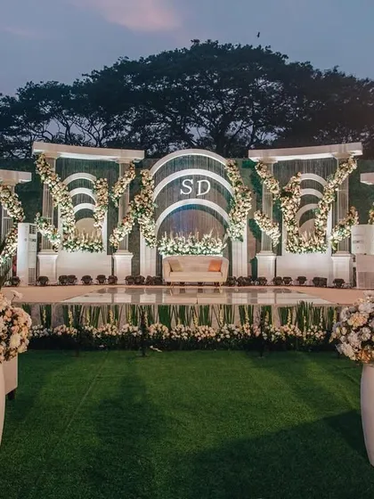 A grand reception stage with neoclassical arches and columns, adorned with cascading white floral arrangements. The monogram in the center adds a personal touch to the majestic design.