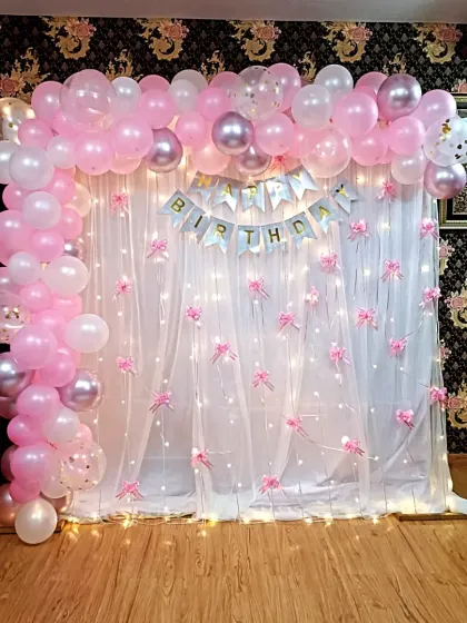 A sweet and simple home decoration for a birthday. A white curtain with fairy lights is framed by a pink and white balloon arch, creating a lovely photo corner.