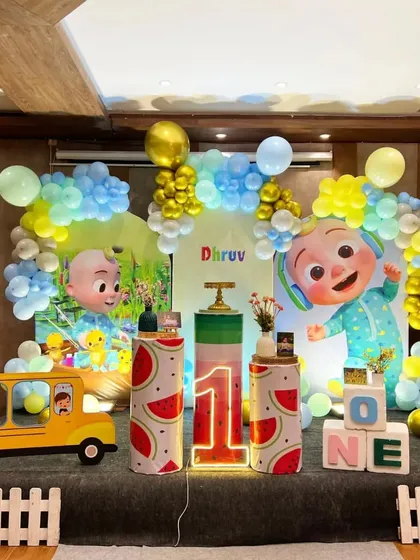 A large-scale Cocomelon stage decoration for a first birthday. This setup features multiple character props, a Cocomelon school bus, themed pedestals, and a light-up number '1'.