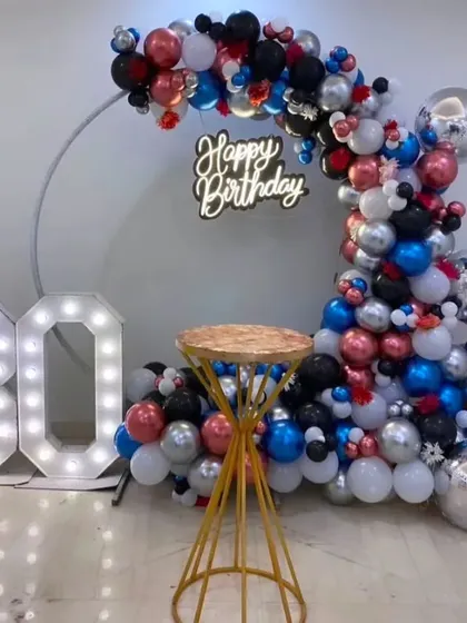 A vibrant 30th birthday party decoration. A circular frame is adorned with a colorful mix of red, blue, black, and silver balloons, complemented by a neon sign and light-up '30' numbers.