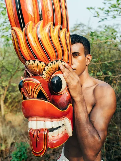 A portrait from the "Nazaar" series, where the model's face is partially obscured by a large, colorful traditional mask. The outdoor setting adds an earthy, raw quality.