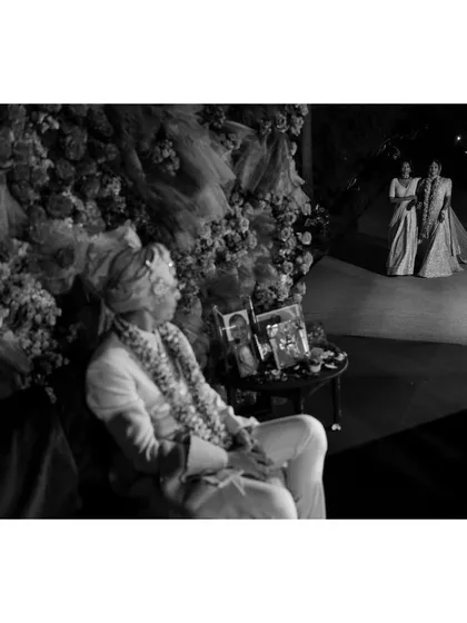 A groom waits patiently for his bride to arrive. This black and white photo uses layers to tell a story, with the groom in the foreground and the bride's arrival in the distance.