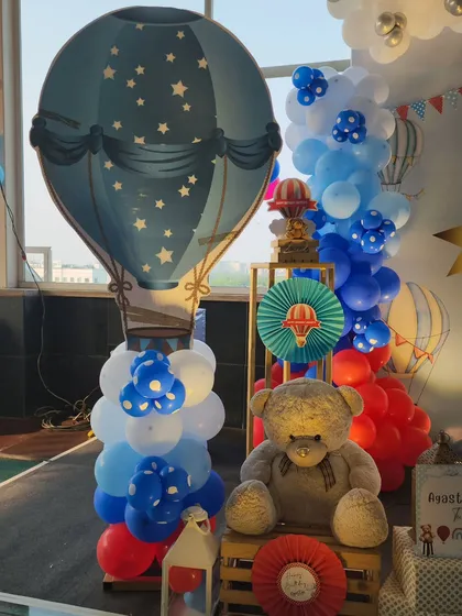 A detailed shot of the decor next to the main stage. It features a large teddy bear, a blue hot air balloon cutout, and a column of blue and white balloons, creating a full and festive look.