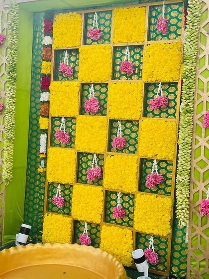 A vibrant Haldi backdrop with a checkerboard pattern of yellow marigolds. The setup includes a traditional brass vessel (urli) for the ceremony, framed by decorative panels.