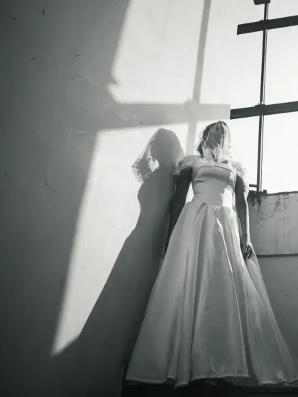 An artistic black and white bridal portrait using shadow and light. The bride stands veiled against a wall, creating a dramatic and mysterious image.