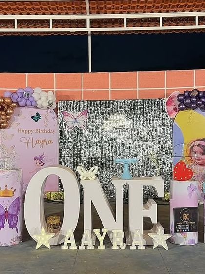 An outdoor butterfly-themed setup for a first birthday. This decoration features large cutout wings, a silver shimmer wall, and personalized plinths, all arranged under a canopy.