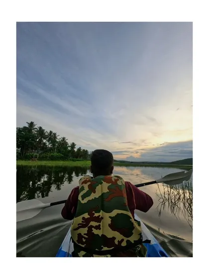 A solo kayaker enjoys the tranquility of the backwaters, surrounded by the reflection of the sky and palm trees. A perfect moment of peace.