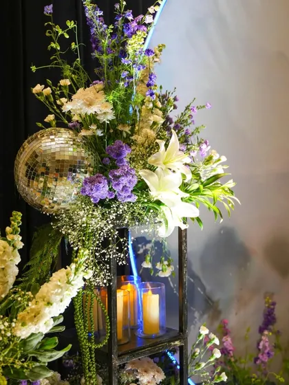 A stunning corner where florals meet glam. A disco ball is nestled within an arrangement of white lilies and purple asters, with candles glowing in a modern black stand.