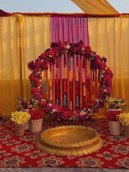A beautiful Haldi setup on a terrace with a floral ring made of red and yellow flowers. The large brass urli in the center is perfect for the ceremony, set against a backdrop of colorful drapes.