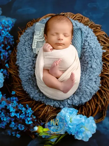 A sleeping newborn swaddled in white, nestled in a basket lined with blue fur and surrounded by blue flowers.