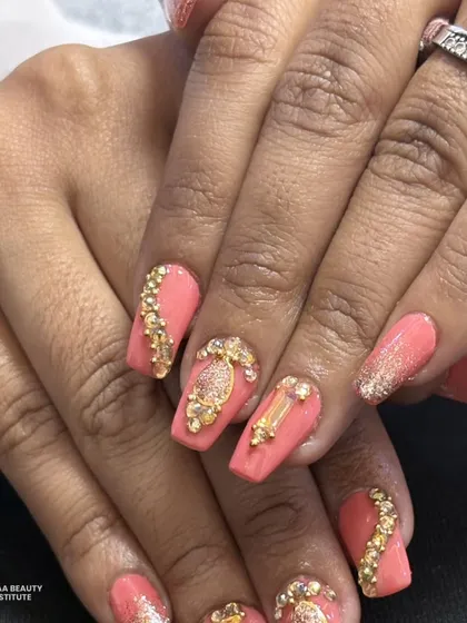 A stunning coral-pink set adorned with intricate gold charms and crystals. The arrangement of the gems creates a luxurious, jewelry-like effect on the nails.