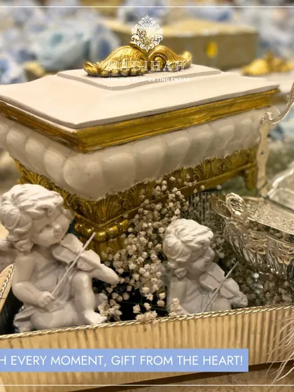 A close-up on the charming details of a baby boy hamper. This shot focuses on the two cherub figurines playing violins, nestled among baby's breath next to an ornate white and gold keepsake box.