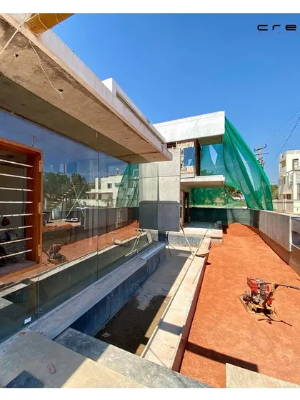 An under-construction view of The Quarry House, showing the large glass panels being installed. The image highlights the exposed concrete structure and the water channel that will become a key landscape feature.
