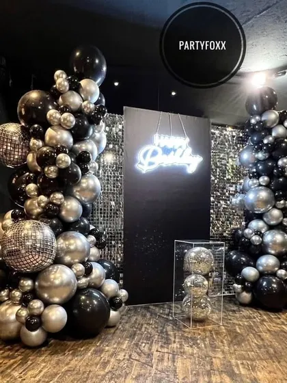 A chic and modern birthday setup with black and silver balloon towers, disco balls, and a neon "Happy Birthday" sign against a sequin backdrop.