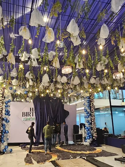 An extravagant birthday party decoration in a large corporate space, featuring a massive ceiling installation with hanging foliage and lights.