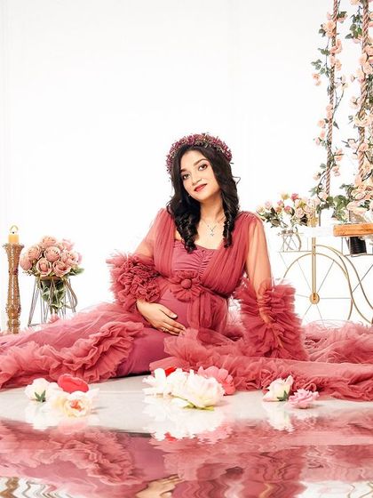 A beautiful solo portrait with a water reflection effect. The mom-to-be is seated, surrounded by the flowing fabric of her dusty rose gown and scattered flowers.