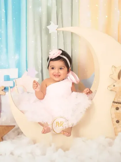 This little girl is sitting on a crescent moon prop, surrounded by stars and clouds, as part of her enchanting first birthday photoshoot.
