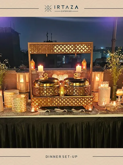 A rooftop dinner setup with two ornate golden chafing dishes as the centerpiece. The lanterns and candles create a magical glow against the night sky, perfect for a special celebration.
