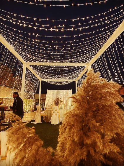 A cozy lounge area for a boho-themed event at night. A canopy of fairy lights illuminates the space, with large pampas grass arrangements adding texture and warmth.