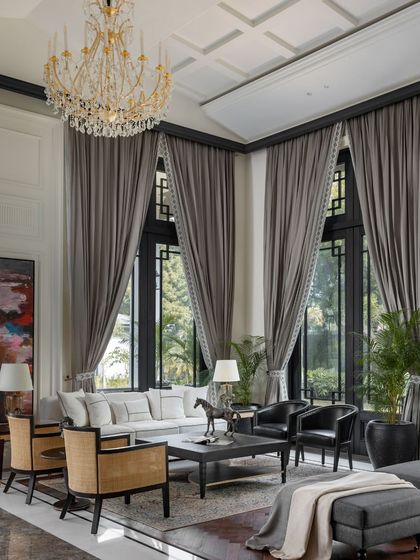Double-height windows in the living room of the House of Verandahs flood the space with natural light and frame views of the surrounding greenery. We used a palette of muted tones and custom furniture to create an atmosphere of both grandeur and comfort.