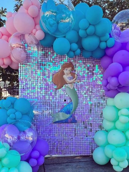 A mermaid-themed birthday setup with an iridescent shimmer wall, character cut-out, and balloon garlands in ocean colors.