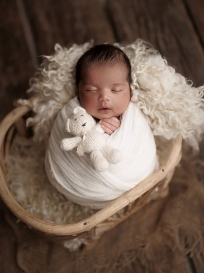 A classic and timeless setup. The baby is wrapped snugly in white and holding a tiny teddy bear, nestled in a rustic basket with soft wool for comfort.