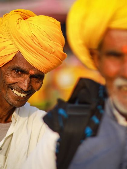 A candid portrait of a smiling man in a yellow turban at a festival. The shallow depth of field separates him from the background, making his joyful expression the focus.