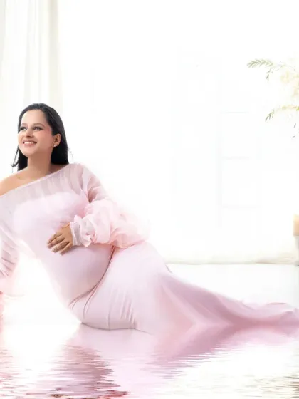The serene beauty of expecting. This bright and airy portrait captures a moment of quiet joy. The mother-to-be, dressed in a soft pink off-the-shoulder gown, is surrounded by gentle light, and the water reflection adds a unique, artistic touch to this peaceful scene.