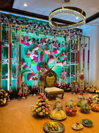 A beautiful and intricate stage setup for a traditional ceremony. The 'tree of life' backdrop, adorned with colorful flowers and surrounded by fruit baskets, creates a vibrant and auspicious setting.