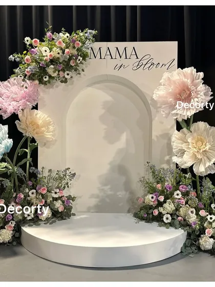 An elegant 'Mama in Bloom' baby shower backdrop. This design uses a clean white arch panel adorned with large, artistic paper flowers and fresh floral arrangements for a sophisticated and modern look.