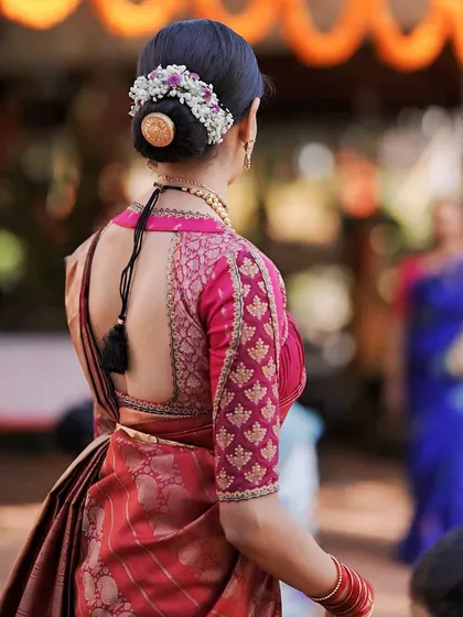 The back view of a pink bridal blouse with a deep U-cut and intricate gold embroidery. The design focuses on creating a beautiful frame for the bride's back, complemented by a simple floral hairstyle.