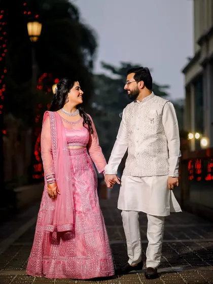 A classic portrait of the couple holding hands and walking. The bride's pink lehenga and the groom's white sherwani create a soft, romantic color palette.