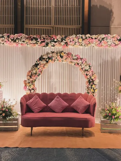 A lovely engagement stage with a circular floral arch in shades of pink. The modern pink sofa and wooden crate flower stands add a rustic-chic touch.