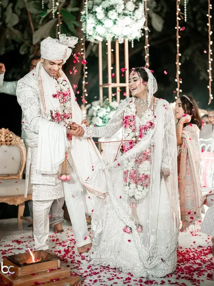 The first steps as husband and wife. The couple takes their pheras together in stunning all-white outfits, surrounded by scattered petals.