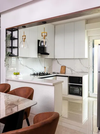 Another angle of the all-white kitchen, showing the open layout and breakfast bar. The warm leather-tone chairs and gold pendant lights add a touch of warmth and prevent the space from feeling too sterile.