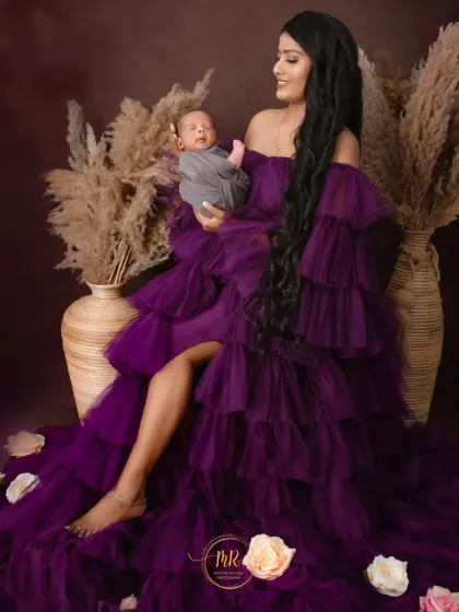 A full-length portrait of a mother in a stunning purple gown, looking lovingly at the newborn in her arms.