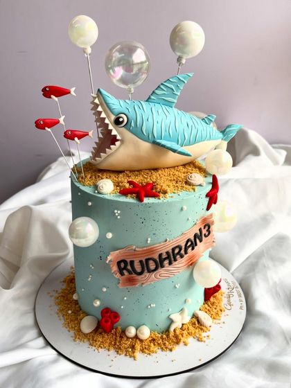 Dive into a sweet ocean adventure with this shark-themed cake. It features a hand-sculpted shark topper, edible sand, red fish, and bubble-like spheres, creating a fun underwater scene for a third birthday.